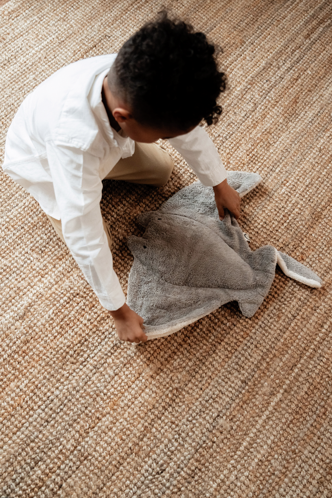A young child with curly hair and a white shirt picks up the Senger Naturwelt Stuffed Animal - Large Manta Ray from a woven, tan-colored rug.