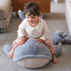 A toddler with short brown hair smiles while sitting on a large, plush Ride-On Whale Pouf on a striped carpet. A smaller stuffed animal rests nearby on the floor.