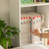 A hand picks a white "Diamond Painting Kit - Positano by Petra Lidze" box from a shelf lined with similar kits, beside a green leafy plant and a white llama figurine.