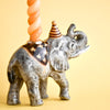 The Elephant Cake Topper is a porcelain figurine with a striped party hat, holding an orange spiral candle on its back, displayed on a plain yellow background.