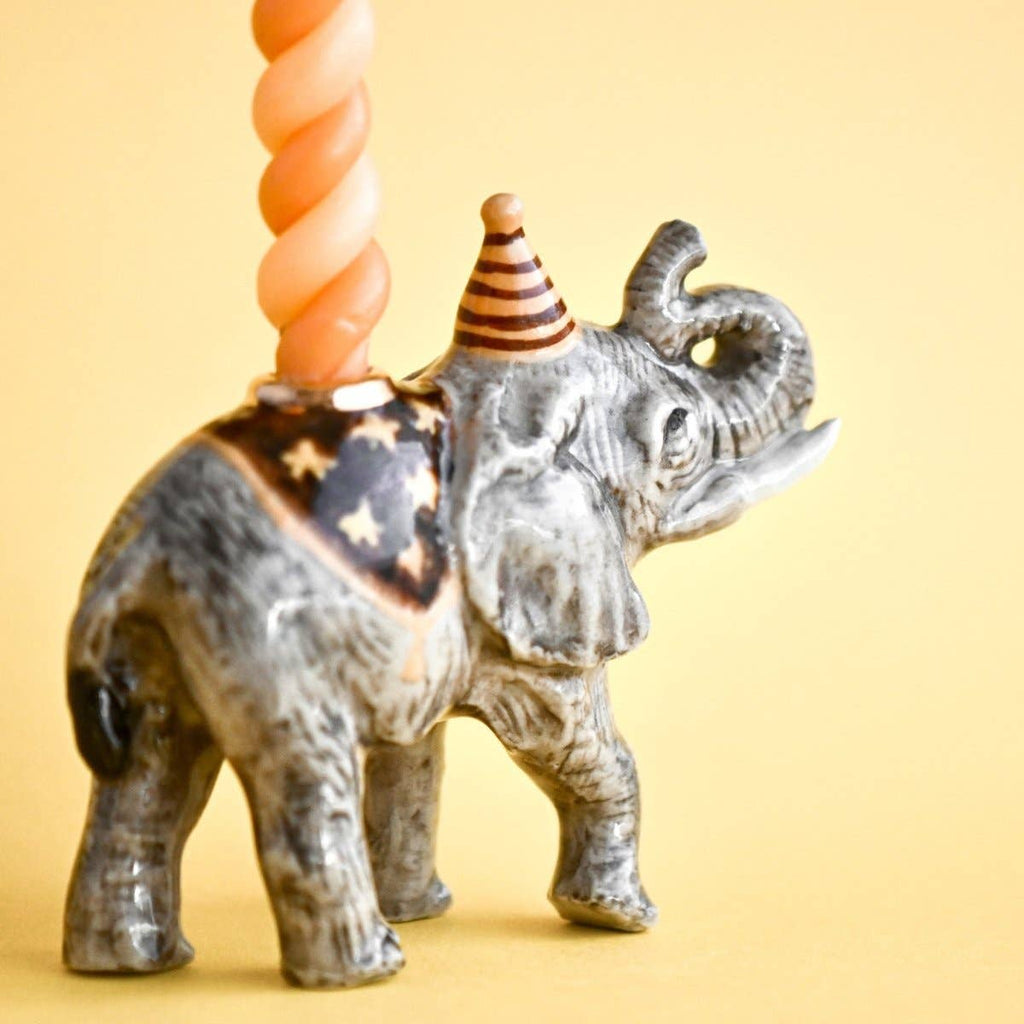 The Elephant Cake Topper is a porcelain figurine with a striped party hat, holding an orange spiral candle on its back, displayed on a plain yellow background.
