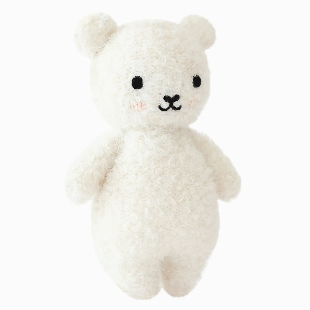 The Cuddle + Kind Baby Bouclé Bear - Ivory is a plush white teddy bear with black eyes, a small black nose and mouth, and pink blush spots on its cheeks, hand-knit from Peruvian alpaca yarn, standing upright against a white background.