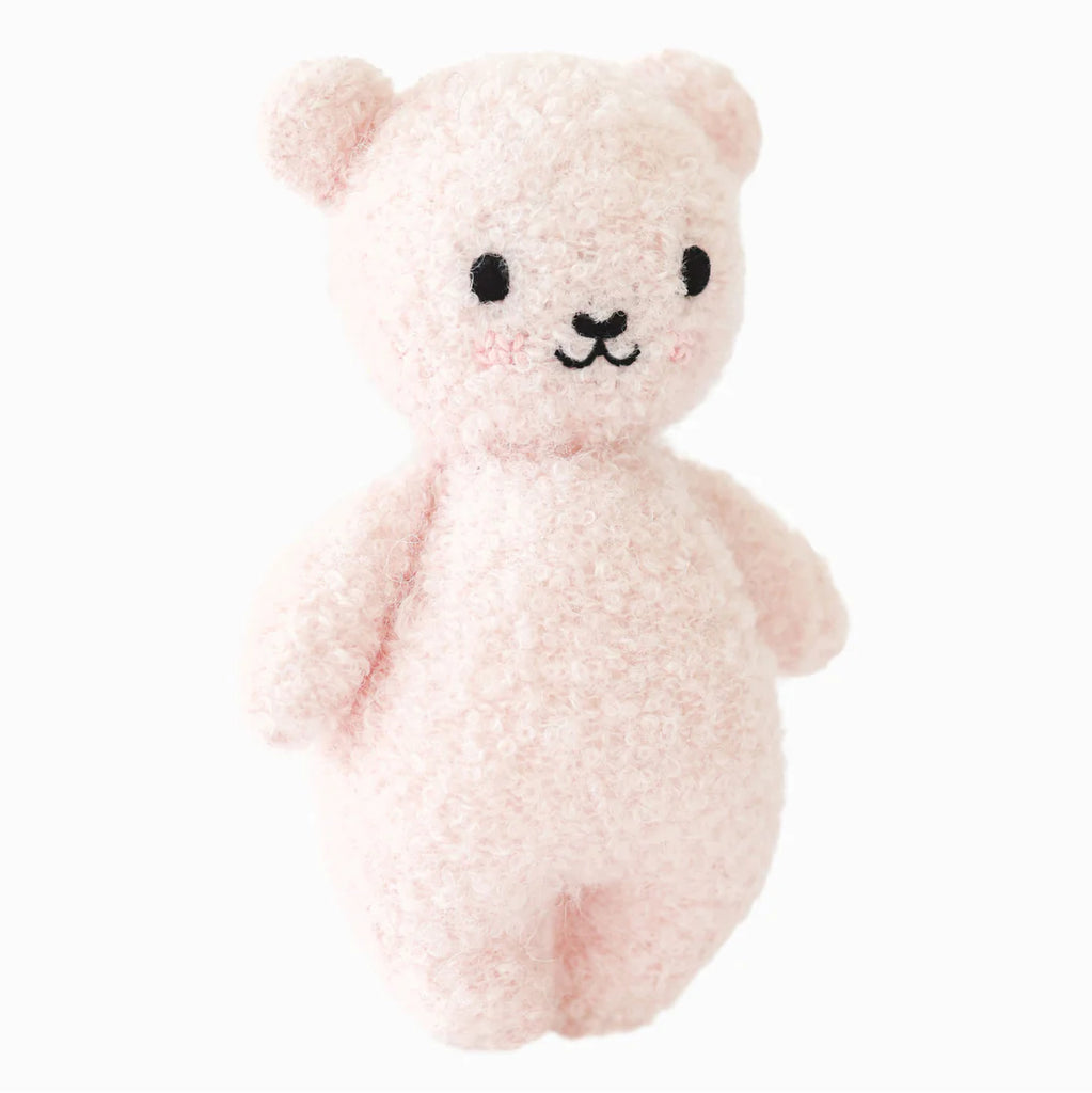 The Cuddle + Kind Baby Bouclé Bear - Strawberry is a soft, light pink plush bear hand-knit with alpaca yarn, featuring round ears, black embroidered facial details, rosy cheeks, and stands upright against a white background.