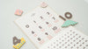 The Magnetic Number Play Set features a minimalist calendar with playful magnets—dinosaur, flower, and numbers 4 and 10—on a light background. Large numbers accented by pink dots add charm, reminiscent of classic magnet puzzles.