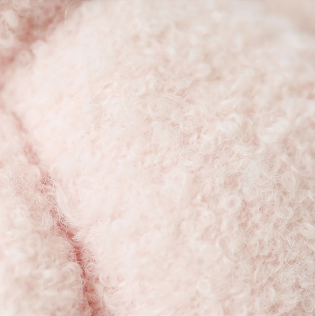 Close-up of the Cuddle + Kind Baby Bouclé Bear - Strawberry, featuring soft, light pink curly bouclé fabric with a fluffy, cozy hand-knit appearance.