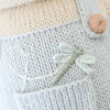 Close-up of light blue knit fabric with a small embroidered dragonfly and white stitched line; a beige button and cream knitting from the fair trade, hand-knit Cuddle + Kind Bailey The Mouse doll appear in the background.