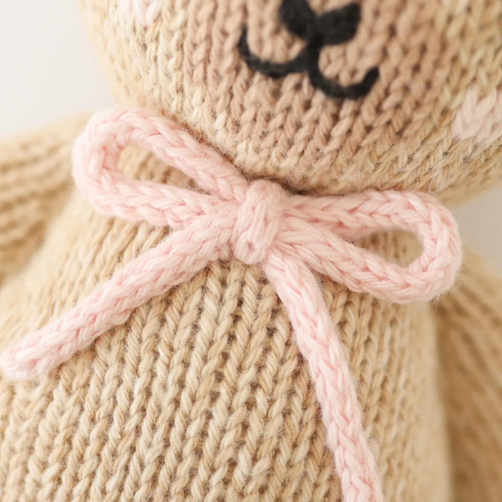 Close-up of Cuddle + Kind Baby Honey Bear - Blush, hand-knit from soft Peruvian cotton yarn with a light pink bow around its neck. Features embroidered black nose and mouth on the teddy bear’s chest and face.