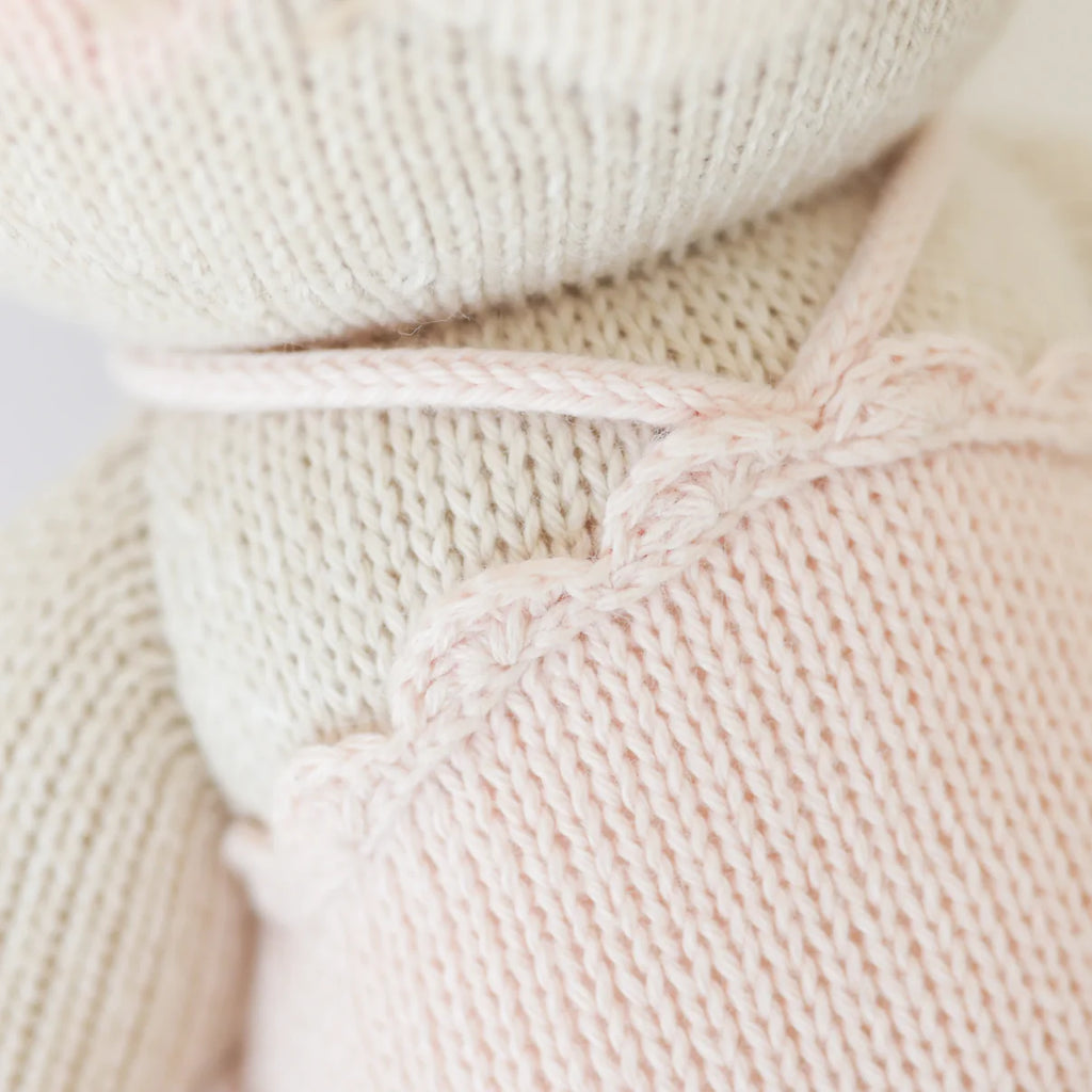 Close-up of Cuddle + Kind Emma The Mouse, a handmade doll knitted from hypoallergenic materials. She wears a light pink outfit with scalloped edges and a white cord necklace, showcasing the yarn’s soft texture and delicate stitching.
