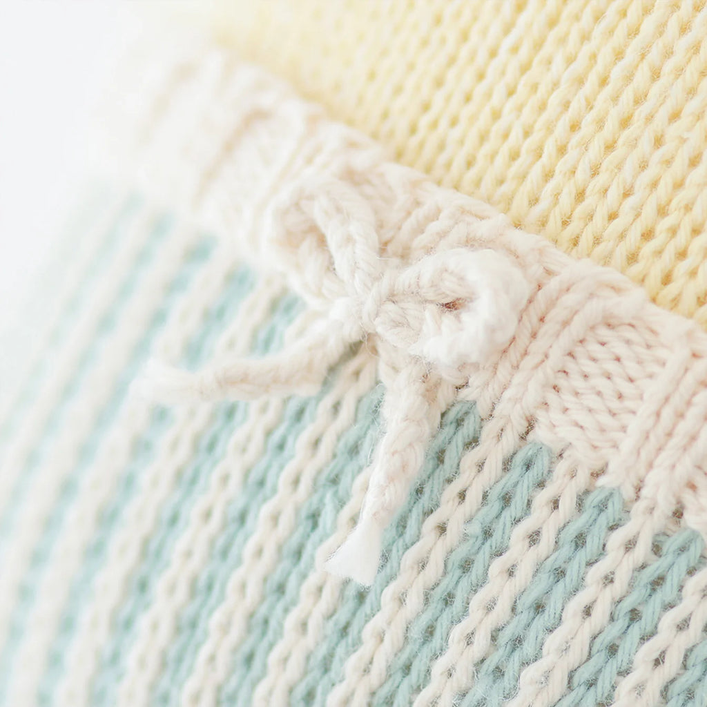 Close-up of the Cuddle + Kind Duckling, featuring soft, hand-knit 100% cotton fabric in pale yellow, blue, and white. A small white bow at the seam highlights its delicate stitching and cozy texture.