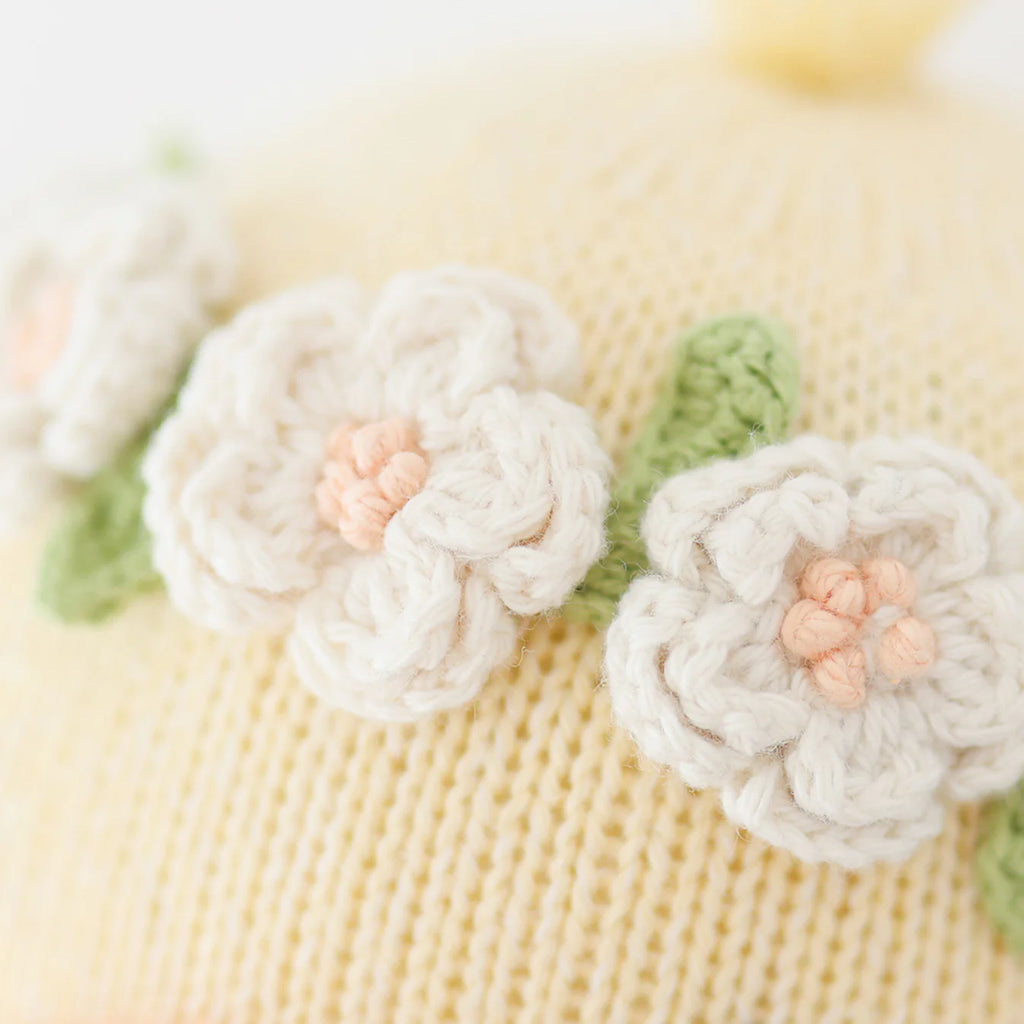 Close-up of the Cuddle + Kind Duckling, a pale yellow knitted fabric made with sustainable fair trade practices, featuring three white crocheted flowers with peach centers and small green leaves.