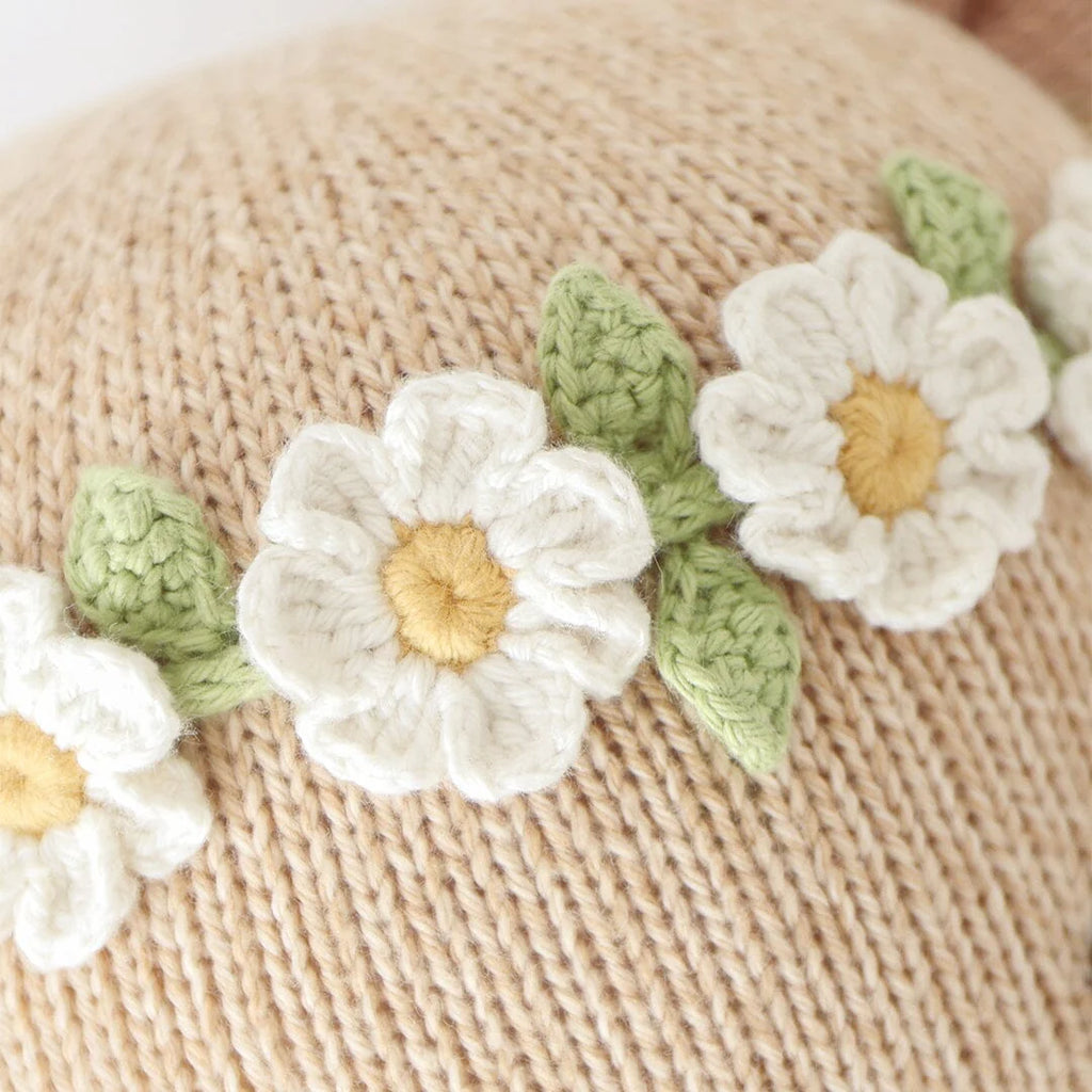 Close-up of Cuddle + Kind Olivia The Honey Bear’s beige hand-knit fabric, decorated with white flowers with yellow centers and green leaves arranged in a row.