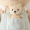 Close-up of Cuddle + Kind Alexander The Honey Bear wearing light blue overalls with a tiny bear in its pocket. Both are hand-knit from light beige yarn, have black stitched eyes and noses, and are stuffed with hypoallergenic polyfill.