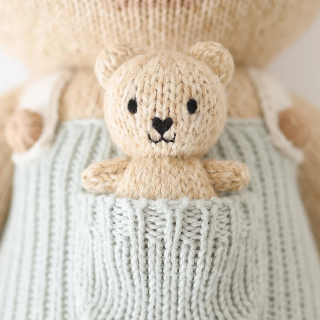 Close-up of Cuddle + Kind Alexander The Honey Bear wearing light blue overalls with a tiny bear in its pocket. Both are hand-knit from light beige yarn, have black stitched eyes and noses, and are stuffed with hypoallergenic polyfill.