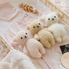 A Cuddle + Kind Baby Bouclé Bear - Honey sits in a wicker basket with soft baby clothes, a wooden rattle, and a quilted blanket, creating a cozy and charming nursery scene.