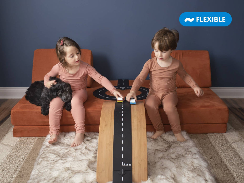 Two young children in matching pajamas play with small toy cars on the WaytoPlay: Medium Flexible Toy Road Set - Expressway between two orange sofas. One child holds a black dog. A “FLEXIBLE” label highlights this imaginative adventure.