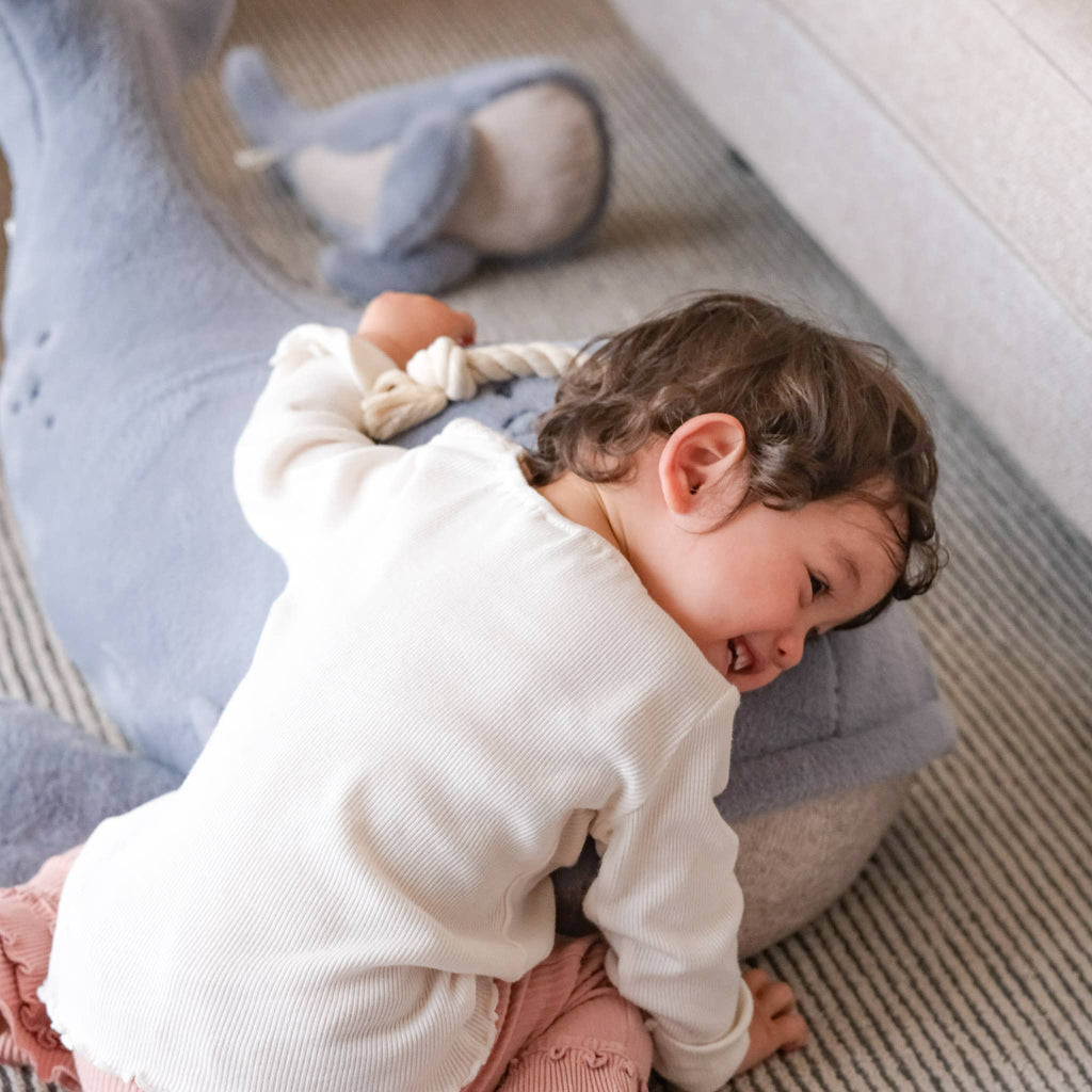 A smiling young child hugs a Ride-On Whale Pouf on a striped carpet, with another plush whale floor cushion in the background.