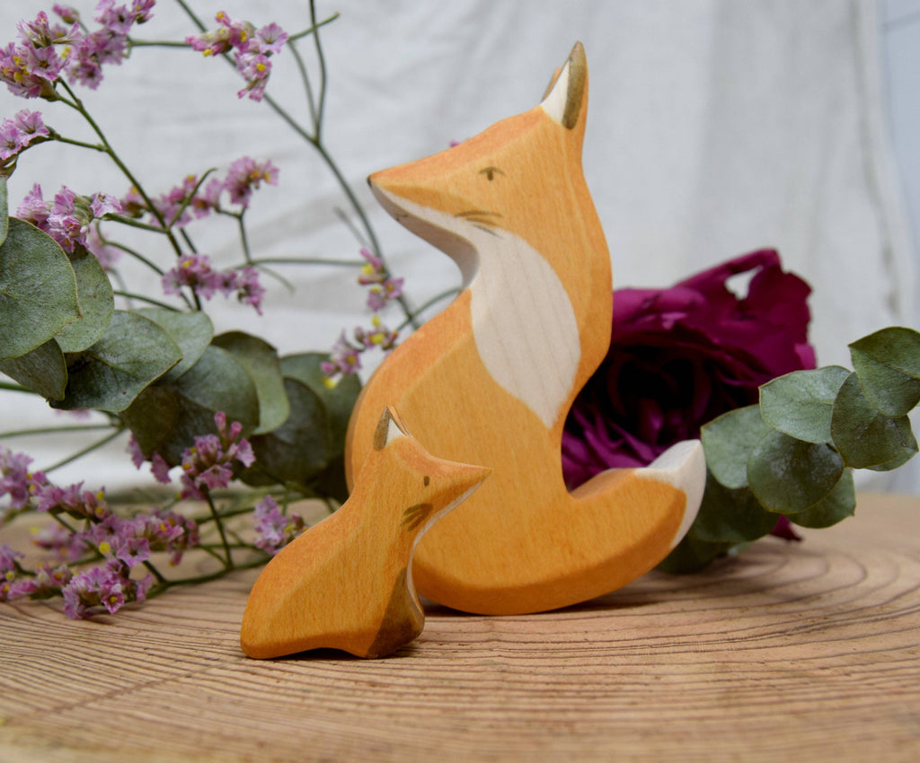 Handmade Wooden Snuggling Foxes—two hand-crafted, sustainable hardwood figurines (one large, one small)—rest on a wooden surface with purple flowers and green leaves, set against a blurred white background.