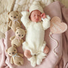 A newborn sleeps on a pink blanket in a fluffy white Peruvian cotton outfit, surrounded by three Cuddle + Kind Baby Honey Bear - Blush plushies and a heart-shaped sign that reads "My First Valentine's Day.