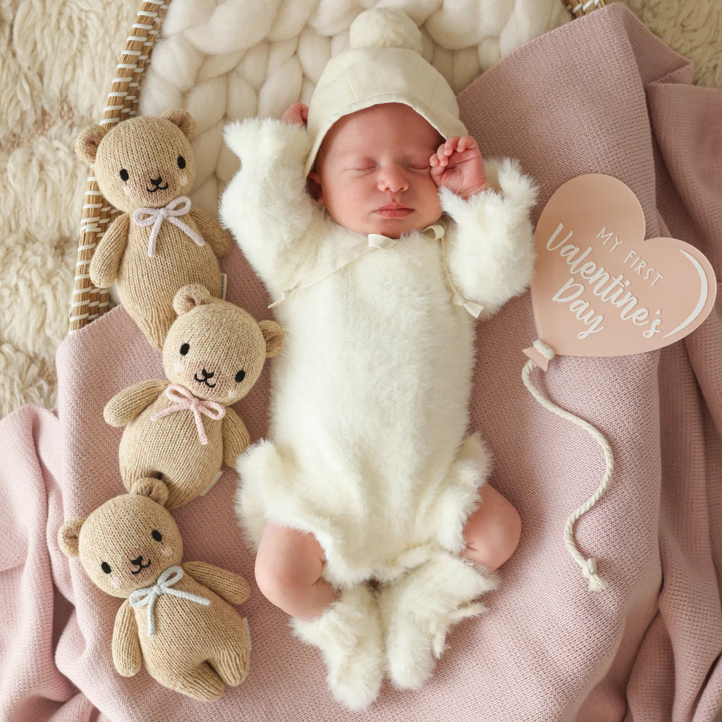 A newborn sleeps on a pink blanket in a fluffy white Peruvian cotton outfit, surrounded by three Cuddle + Kind Baby Honey Bear - Blush plushies and a heart-shaped sign that reads "My First Valentine's Day.