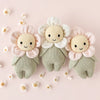 The Cuddle + Kind Baby Daisy - Blush Pink is a hand-knit doll made in Peru from soft Peruvian cotton yarn, featuring a sweet smile, pastel blush petals, and a green body, surrounded by tiny white flowers on a pale pink background.