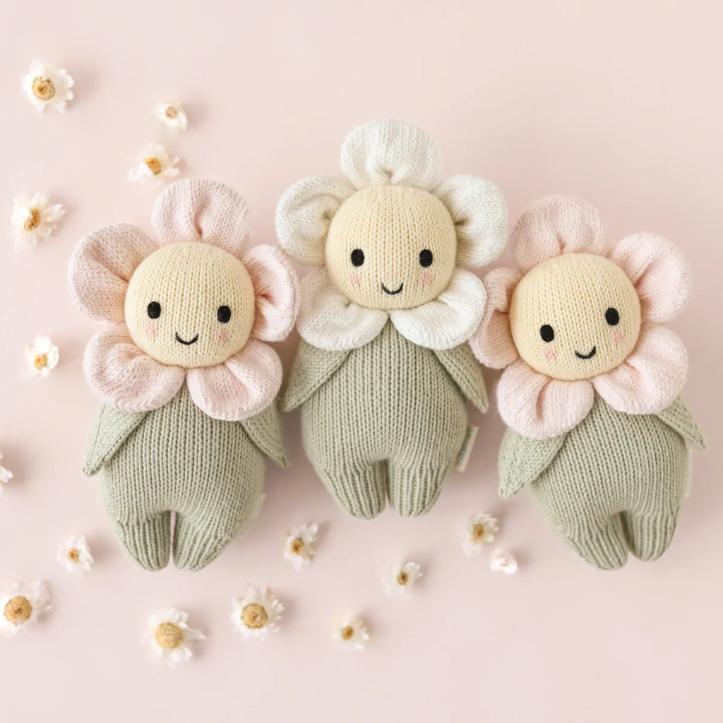 The Cuddle + Kind Baby Daisy - Blush Pink is a hand-knit doll made in Peru from soft Peruvian cotton yarn, featuring a sweet smile, pastel blush petals, and a green body, surrounded by tiny white flowers on a pale pink background.