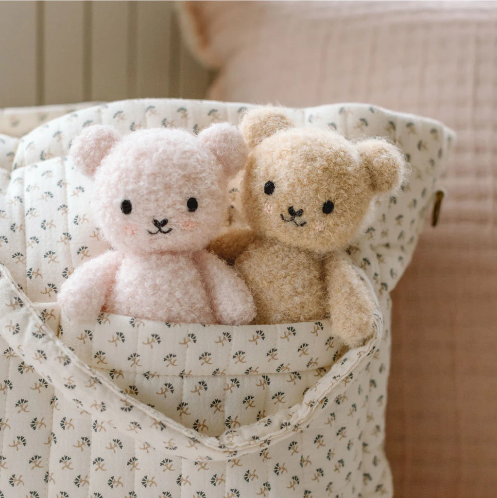 The Cuddle + Kind Baby Bouclé Bear - Honey, crafted with sustainable fair trade practices, sits in a patterned fabric bag next to a soft pink bear, resting against a pale pink textured cushion.