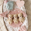 A pink baby romper, mint bonnet, pink booties, and the Cuddle + Kind Baby Honey Bear in Robin’s Egg Blue are arranged on a white blanket beside a heart-shaped "My First Valentine's Day" sign.