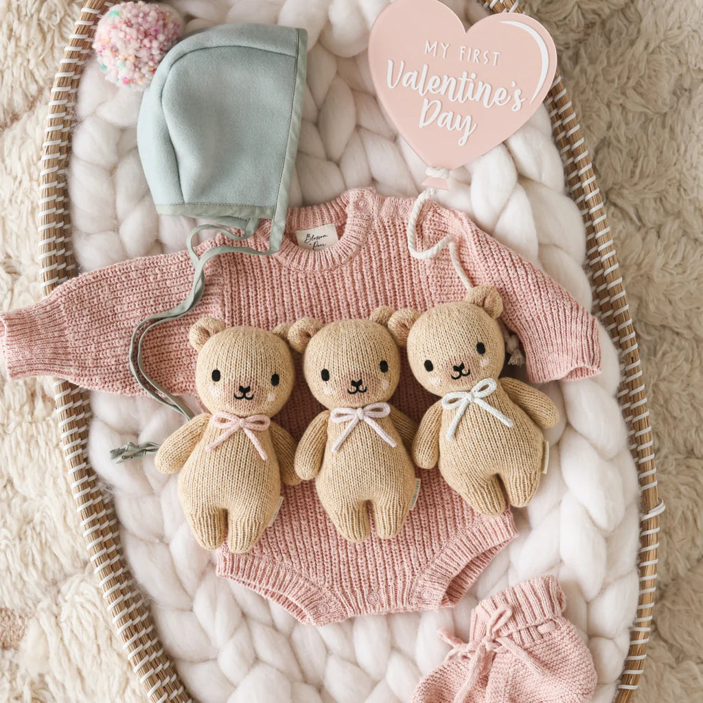 A pink knitted baby romper, mint bonnet, pink knit pants, and three Cuddle + Kind Baby Honey Bear - Blush toys with bows are displayed on a chunky white blanket with a heart sign reading "My First Valentine’s Day." Some pieces are handmade in Peru.