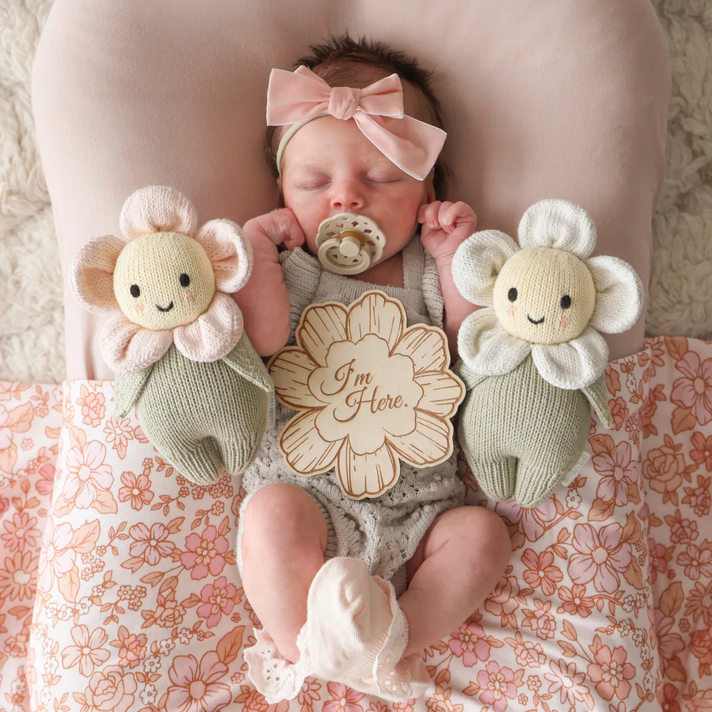 A sleeping baby with a pink bow headband and pacifier lies on a floral blanket, cuddling the Cuddle + Kind Baby Daisy - Blush Pink, made from natural Peruvian cotton yarn, and holds a wooden sign that reads “I’m Here.”.