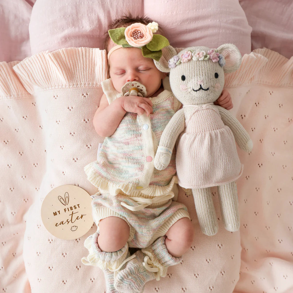 A newborn baby in a flower headband sleeps on a pink blanket, holding the Cuddle + Kind Emma The Mouse doll in a pink skirt. A wooden plaque beside the baby reads “My First Easter.”.