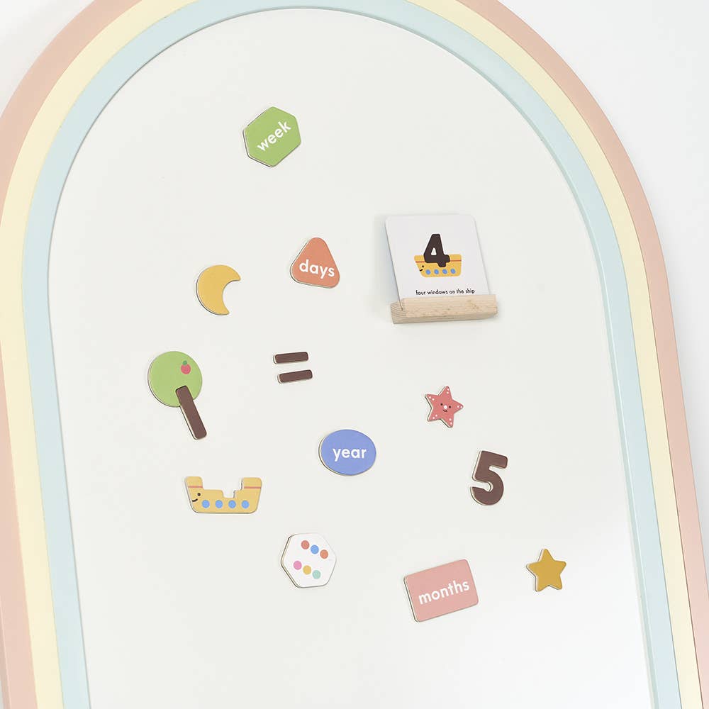 The Magnetic Number Play Set features a whiteboard with a pastel rainbow border, colorful magnetic shapes, number and word magnets like "week," "days," and "months," plus fun symbols such as a tree, star, and equal sign.