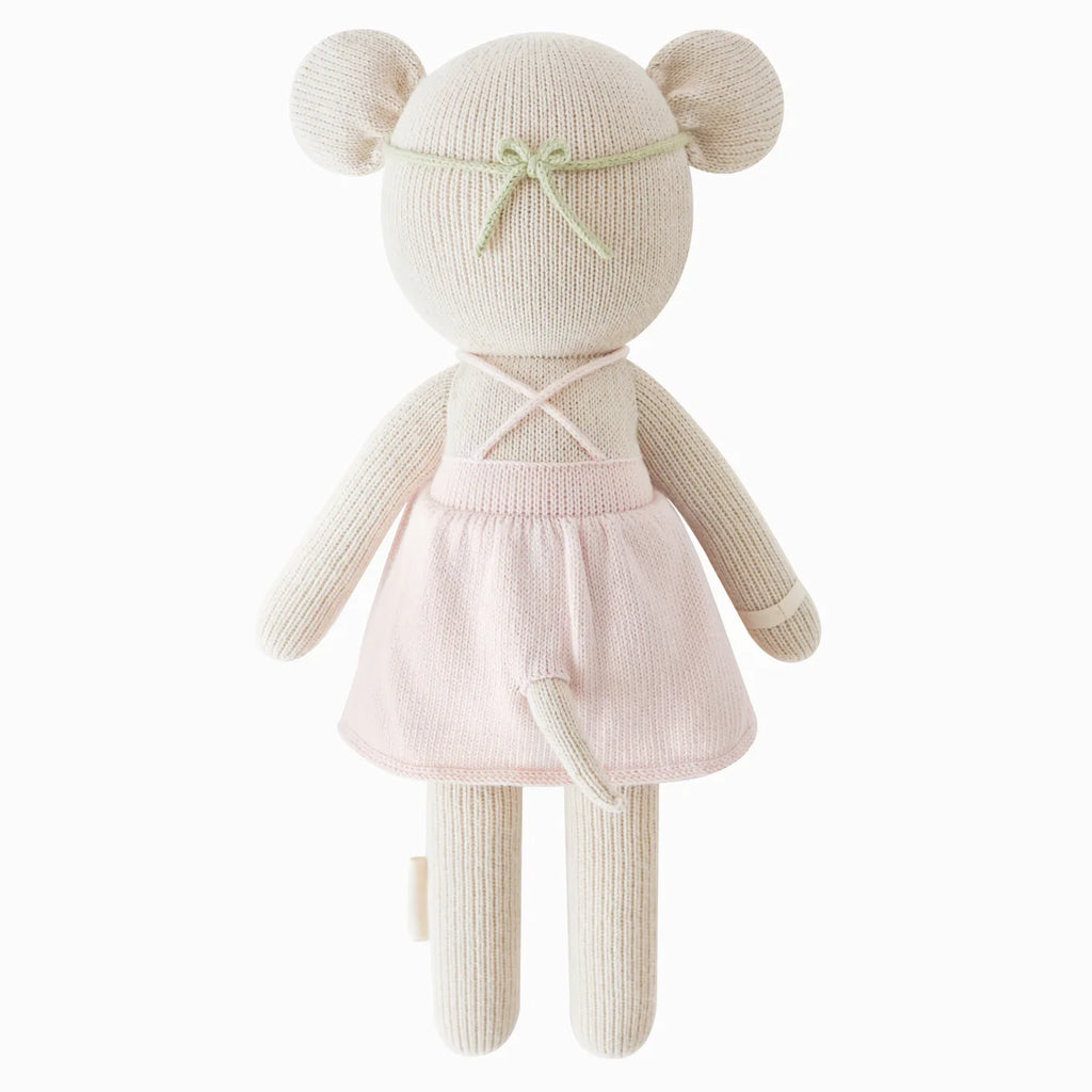 Cuddle + Kind Emma The Mouse is a handmade, hypoallergenic knit stuffed animal resembling a mouse, dressed in a light pink dress with crisscross straps and a pale green bow on its head, shown from the back.