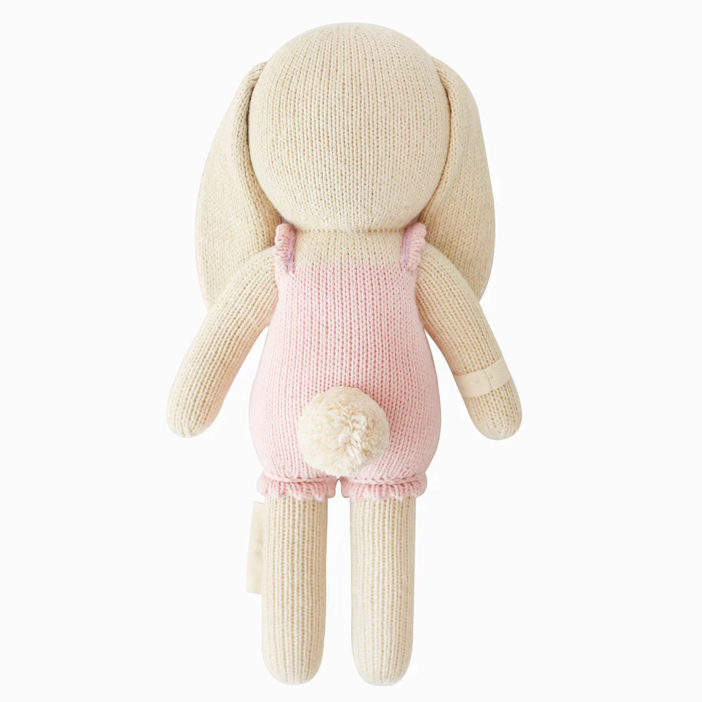 Cuddle + Kind Briar The Bunny - Peony Pink is a hand-knit bunny doll with long ears, a fluffy white tail, and a light pink romper, shown from the back against a white background.