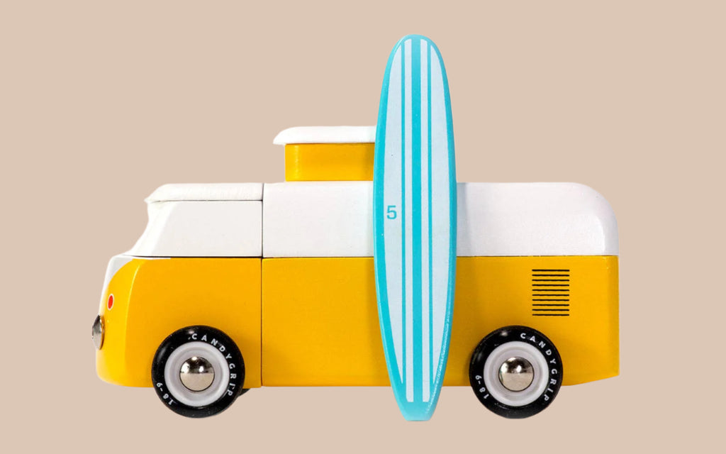 The VW Beach Bus Sunset toy is shown in profile with a magnetic camper top, black wheels, and a blue-striped surfboard leaning against its side on a plain beige background.