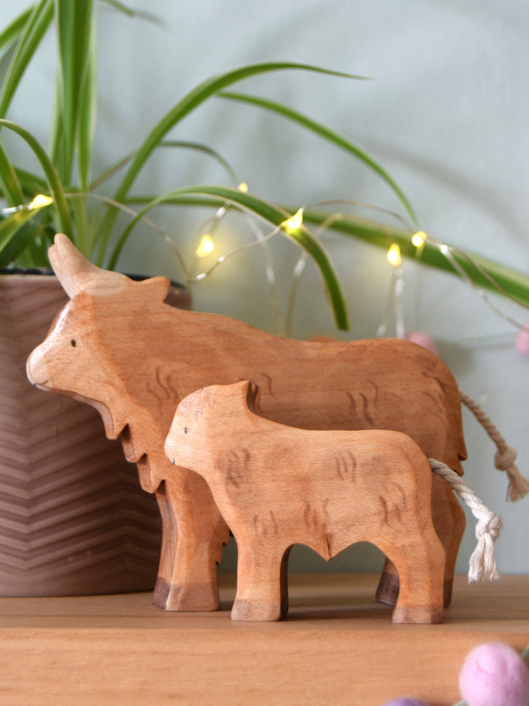 A Handmade Wooden Highland Calf, crafted from sustainable hardwood and finished with plant-based stain, adds cozy charm to a wooden surface with string lights and a potted plant in the background.
