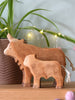 A Handmade Wooden Highland Cow stands on a wooden surface before a potted green plant, with soft fairy lights and pink decorations glowing in the background, accentuating its sustainable hardwood charm.