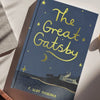 A blue and yellow illustrated hardcover of The Great Gatsby | Collector's Edition rests on a white surface, featuring handwritten-style gold lettering, stars, a crescent moon, and a house by the water.