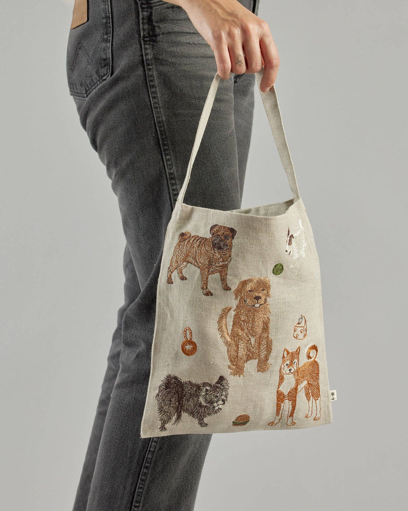 A person holds the "Dogs and Toys Tote" featuring illustrations of various breeds, like a pug, Shih Tzu, and chow chow. This playful tote pairs effortlessly with their dark jeans against a gray background, perfect for pet accessories enthusiasts.