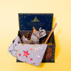 The Elephant Cake Topper features a small toy elephant with a crown and cape, nestled under a star-patterned blanket in a decorative box with a yellow background. The collectible set includes a brown stick and a blue card with gold stars inside the box.