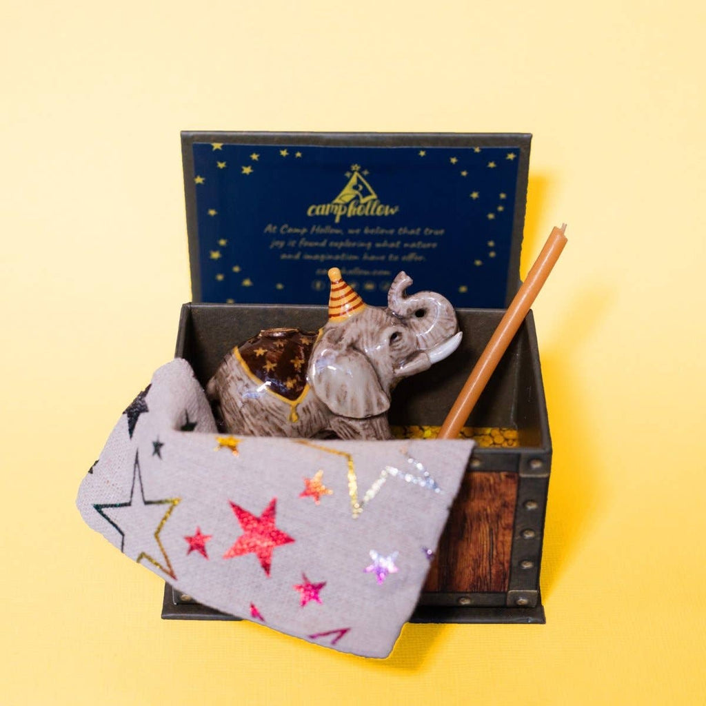 The Elephant Cake Topper features a small toy elephant with a crown and cape, nestled under a star-patterned blanket in a decorative box with a yellow background. The collectible set includes a brown stick and a blue card with gold stars inside the box.