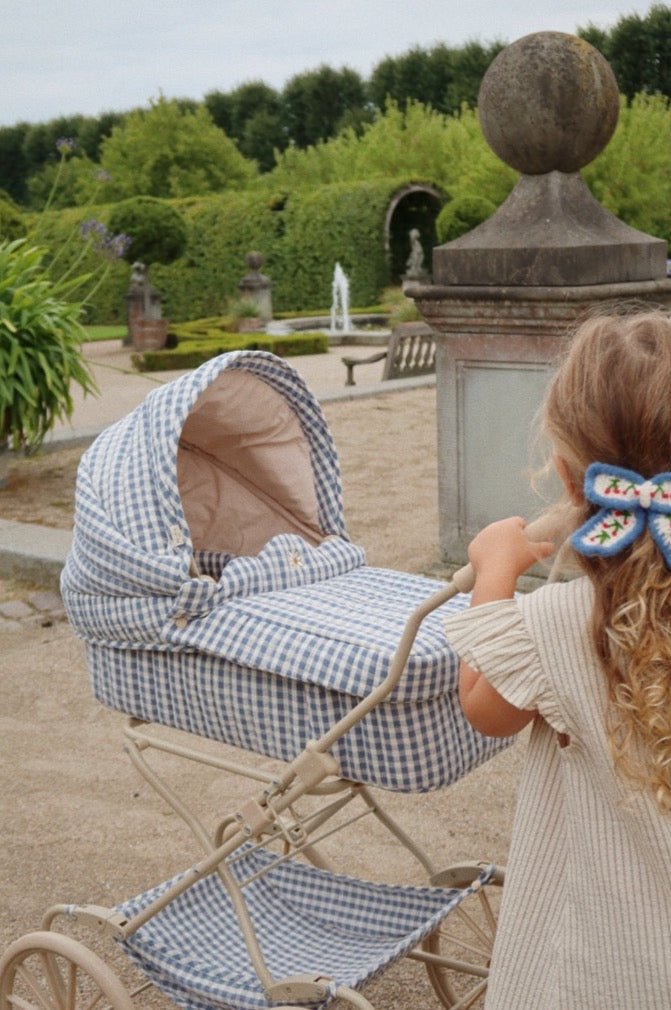 A young girl with blonde hair tied in a ponytail, pushing a Doll Pram - Blue Checkered in a formal garden with topiaries and a fountain in the background.