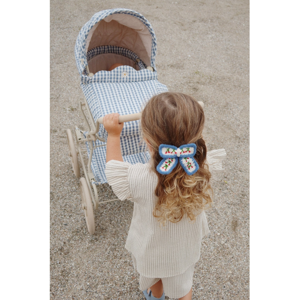A young child with curly blonde hair and a colorful bow stands behind a Doll Pram - Blue Checkered on a gravel surface, viewed from above.