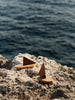 The Ferm Living Opty Boat - Set of 2 features small decorative boats made from FSC-certified oak with brown triangular sails, displayed on a rocky shore with the ocean softly blurred in the background.