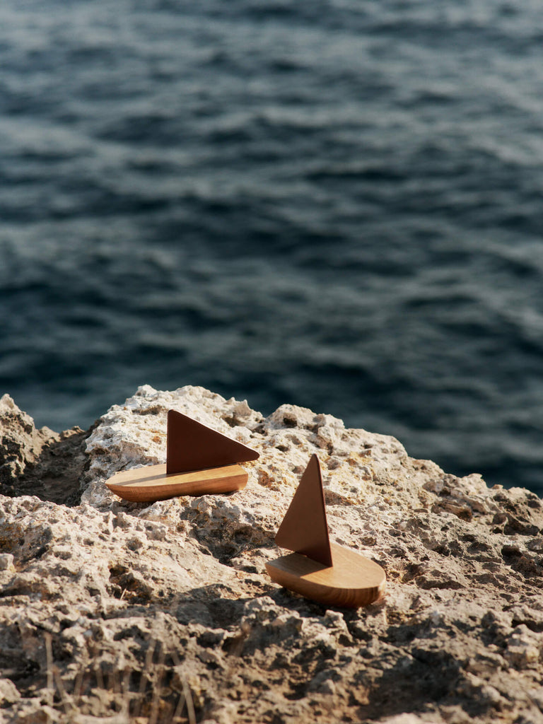 The Ferm Living Opty Boat - Set of 2 features small decorative boats made from FSC-certified oak with brown triangular sails, displayed on a rocky shore with the ocean softly blurred in the background.