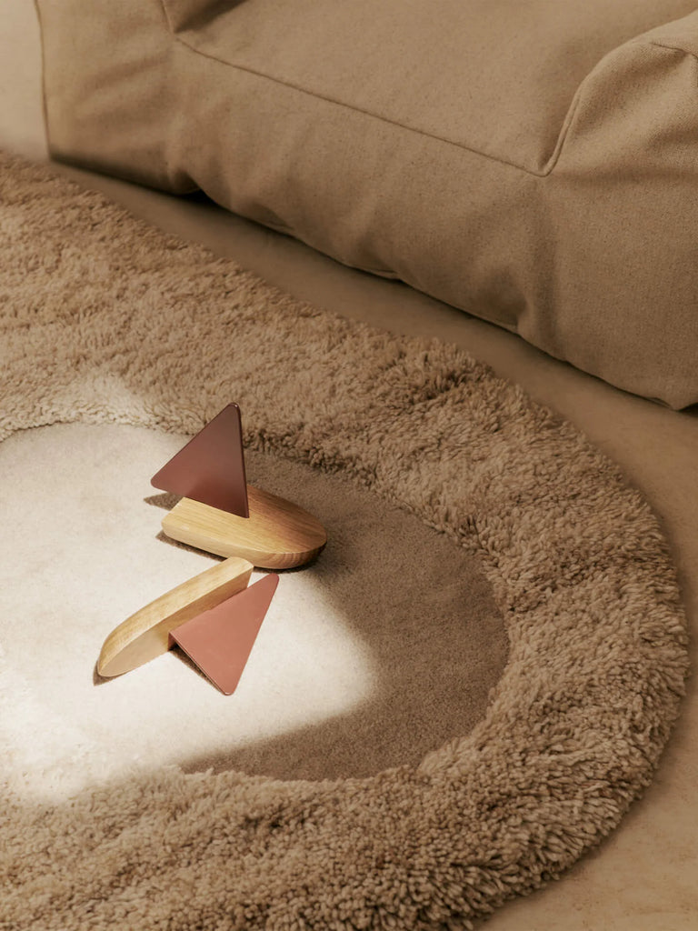 The Ferm Living Opty Boat - Set of 2, crafted from FSC certified oak with pink geometric accents, sits on a round beige rug beside a soft, light couch. Sunlight streams in, adding warmth and coziness to the scene.