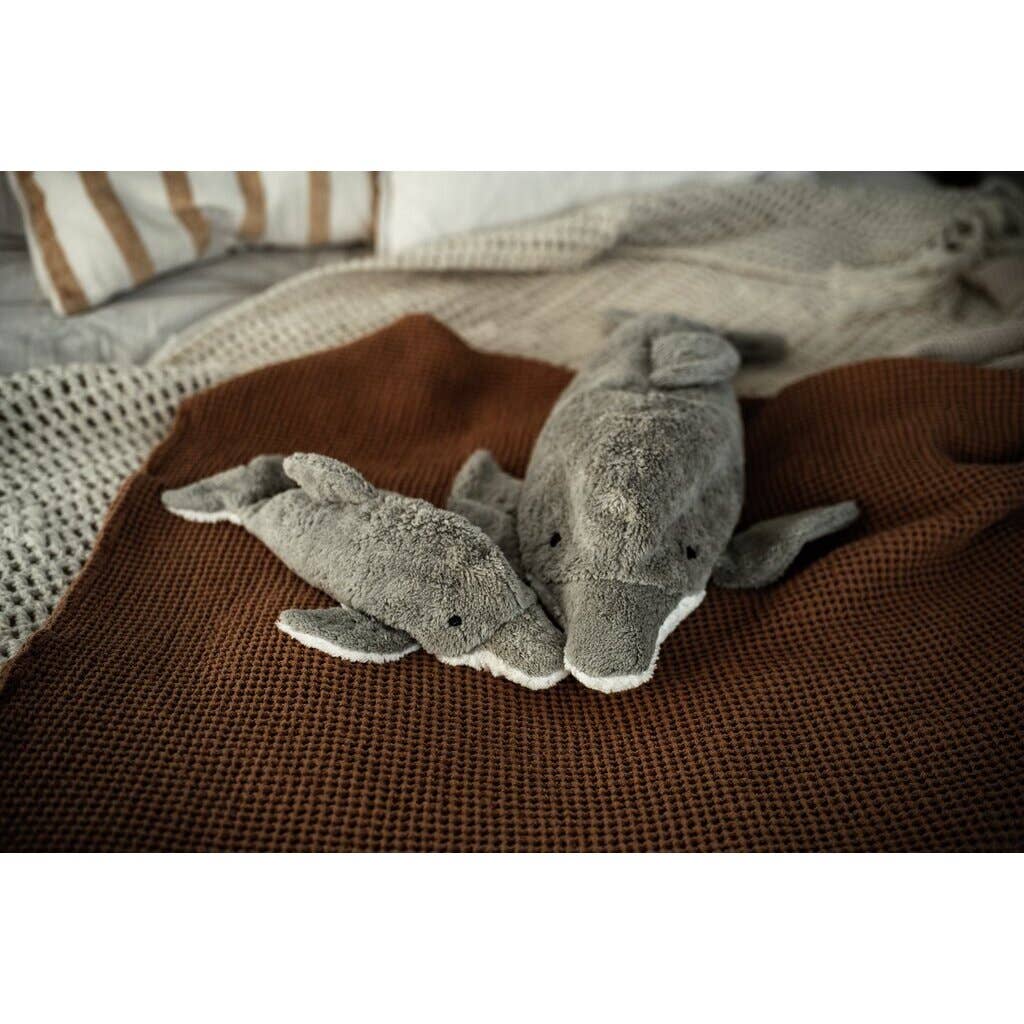 Two Senger Naturwelt Cuddly Animal - Dolphin toys made from sustainable materials rest side by side on a brown textured blanket, with neutral knit and striped bedding in the background.