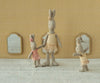 Three Maileg Rabbit, Micro - Ballet Suit And Skirt, Rose dolls stand on a textured surface before a patterned yellow wall with three small mirrors, resembling ballet dancers playfully dancing together in pastel tulle skirts.