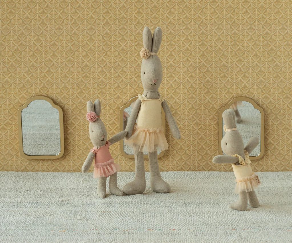 Three Maileg Rabbit, Micro - Ballet Suit And Skirt, Rose dolls stand on a textured surface before a patterned yellow wall with three small mirrors, resembling ballet dancers playfully dancing together in pastel tulle skirts.
