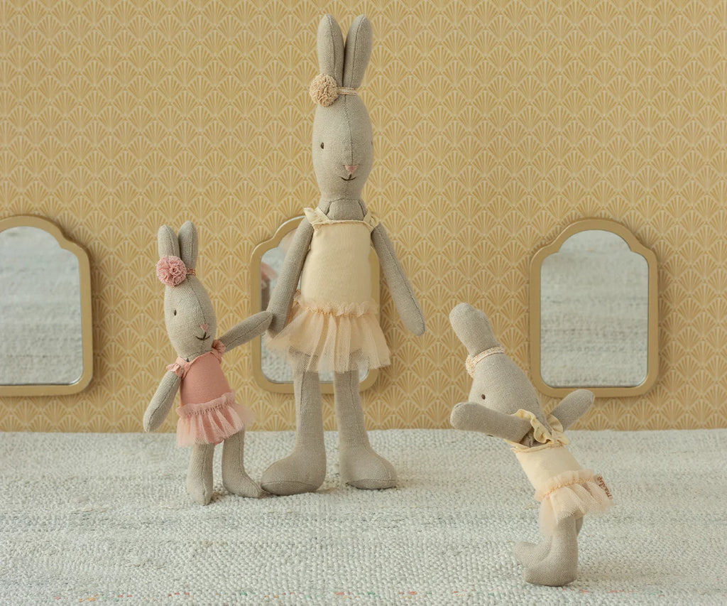 Three Maileg Rabbit, Micro - Ballet Suit and Skirt, Cream stand on a textured surface. Set against yellow patterned wallpaper and three small mirrors, the rabbits are posed like playful ballet dancers in pastel skirts.
