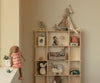 A wooden shelf displays soft toys made from natural fabrics, including rabbits, a penguin, and a giraffe. On the left edge sits the Maileg Bunny, Size 3 - Chocolate Brown - Dress in a softly lit, beige-colored room.